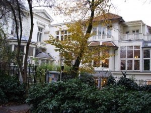 Photo through trees of a 3-story white, L-shaped home with lots of windows